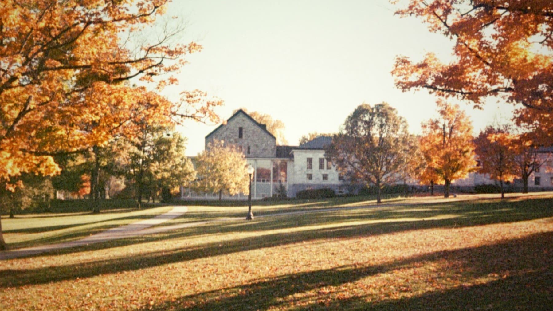 Golden autumn lawn with long shadows and a stone campus hall framed by orange maples.