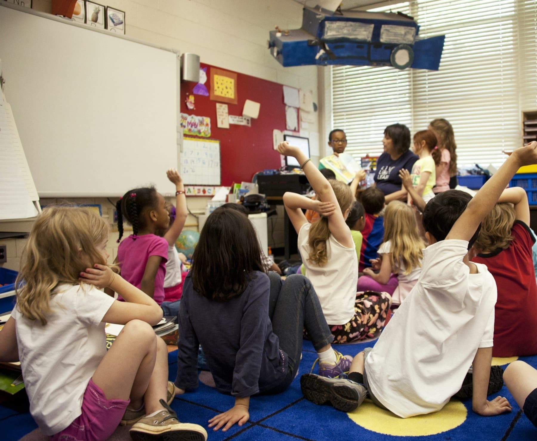 Young students sit on a classroom rug, raising their hands to answer questions during story time