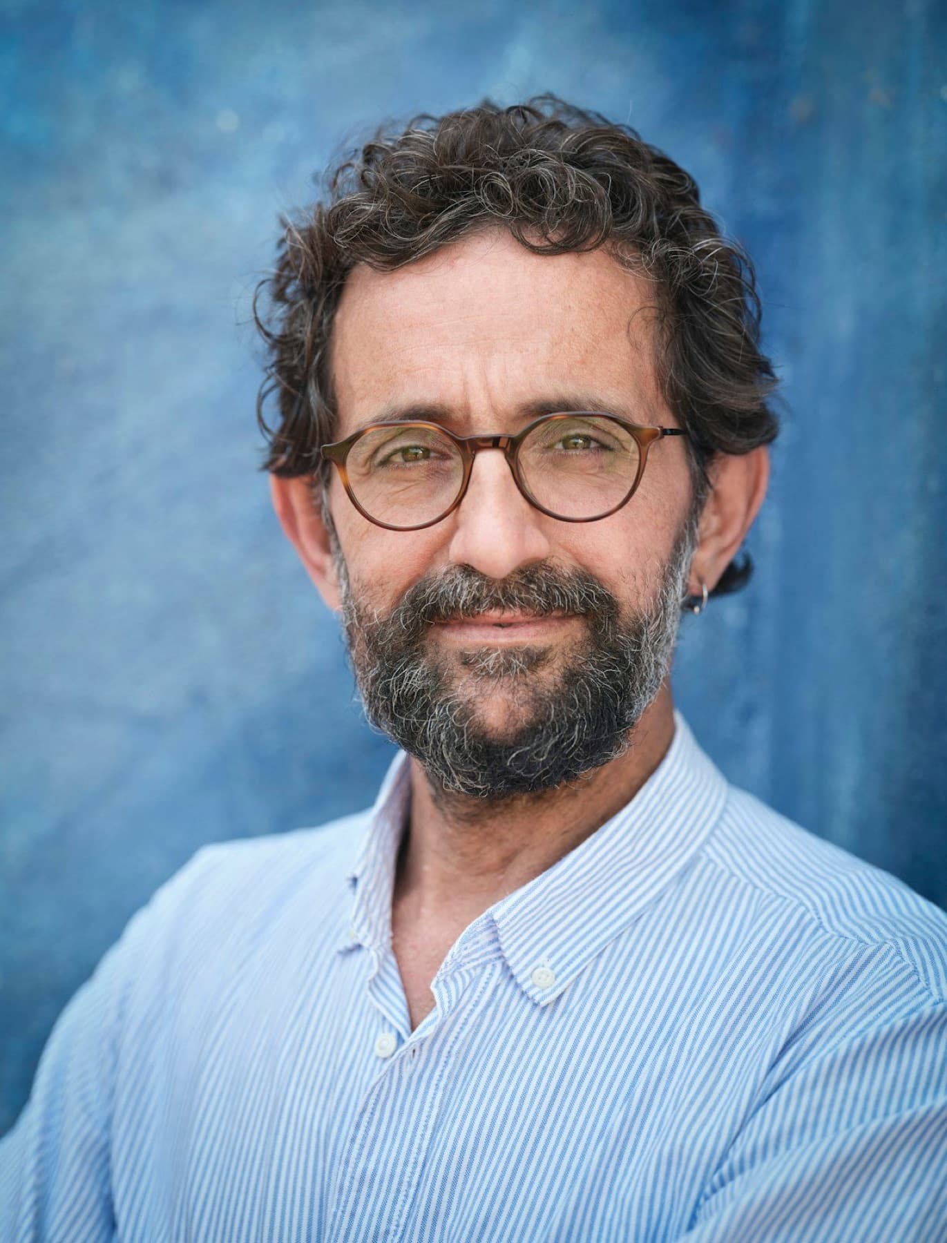 Bearded professor with curly graying hair and round glasses, smiling slightly against a weathered blue wall