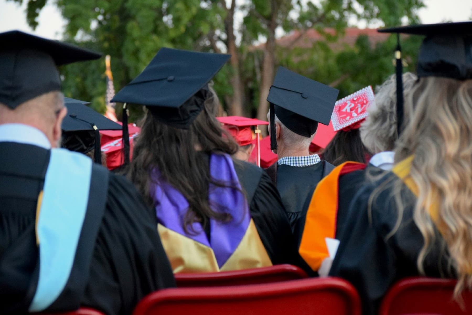 Graduates seen from behind in black caps and colorful academic hoods, seated outdoors at commencement