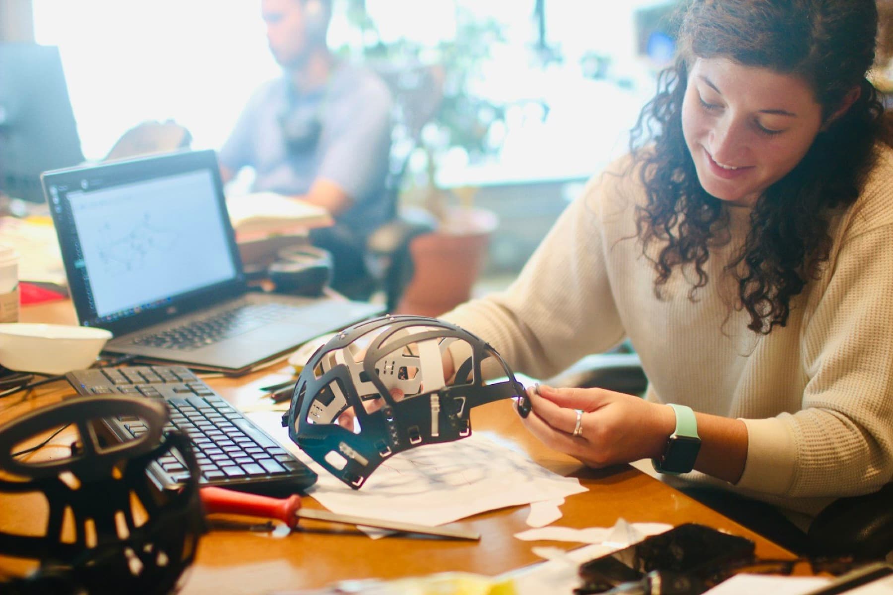 Student smiling as she inspects a 3D-printed prototype part at a cluttered engineering workstation