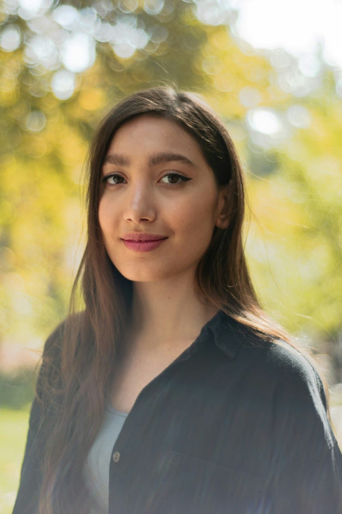 Student with long dark hair smiling softly outdoors, sunlit autumn trees blurred behind her