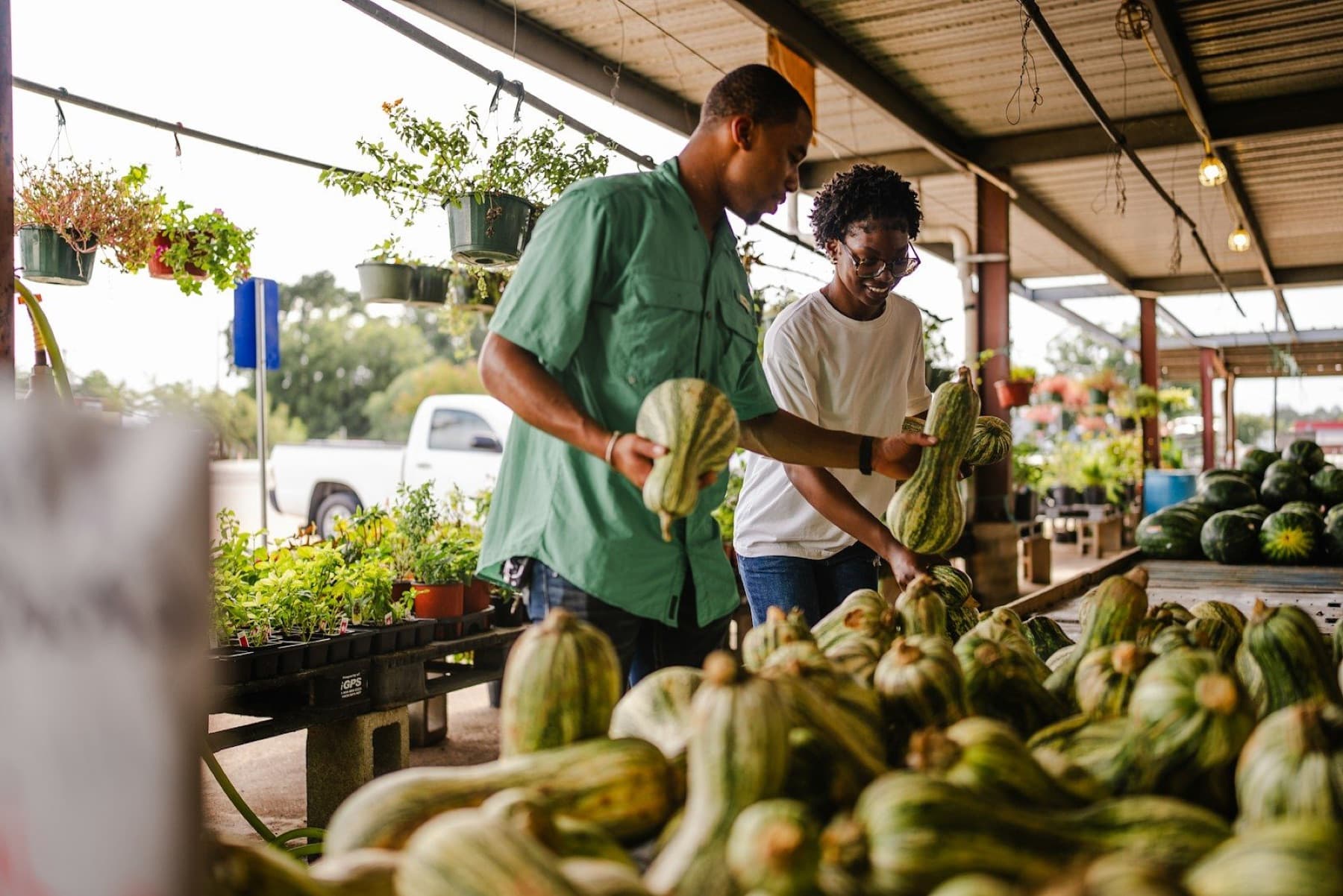 Two young men smile as they pick out striped squash at a covered farmers market stall