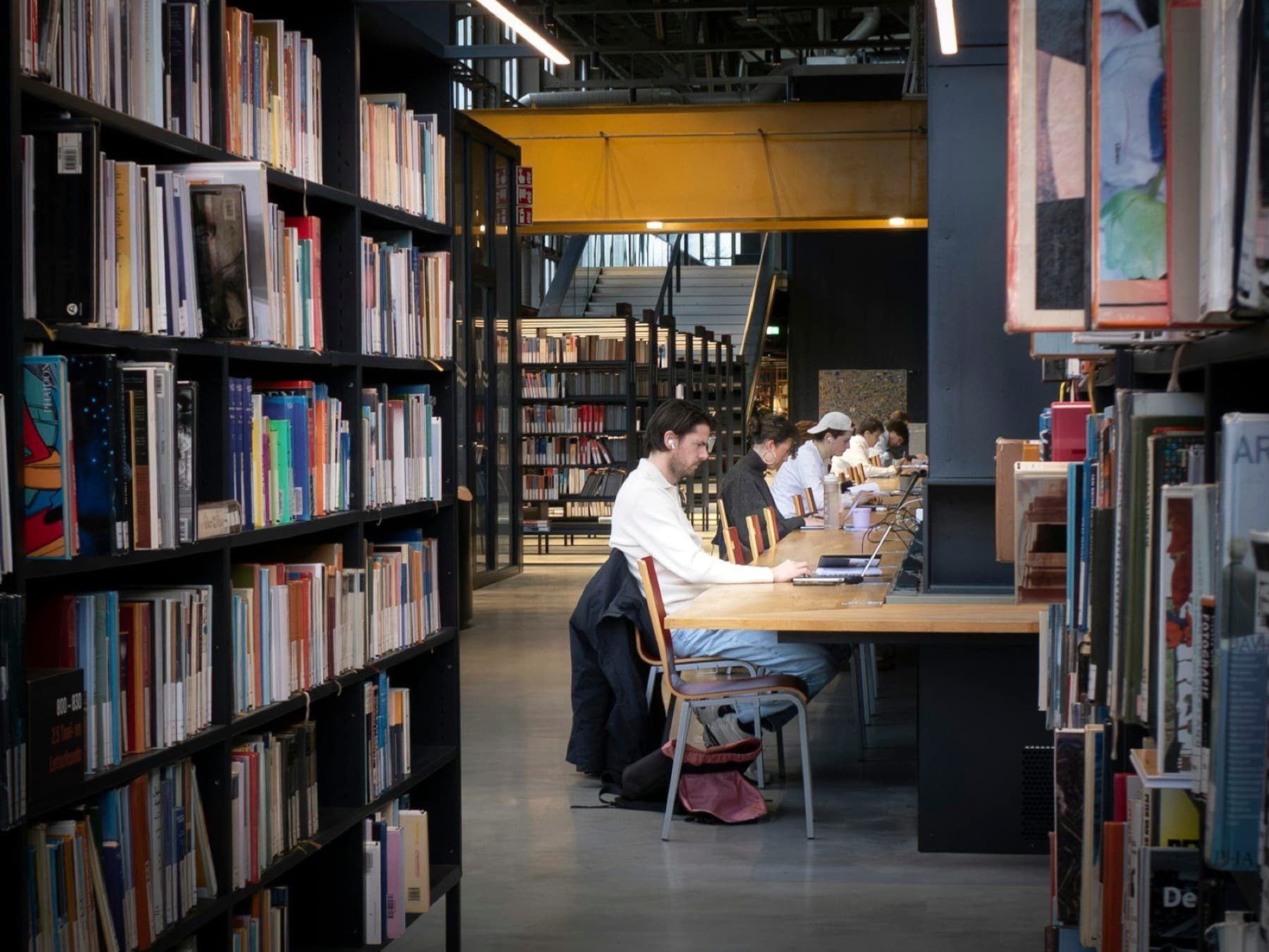 People read and work on laptops at a long wooden table between tall shelves of books