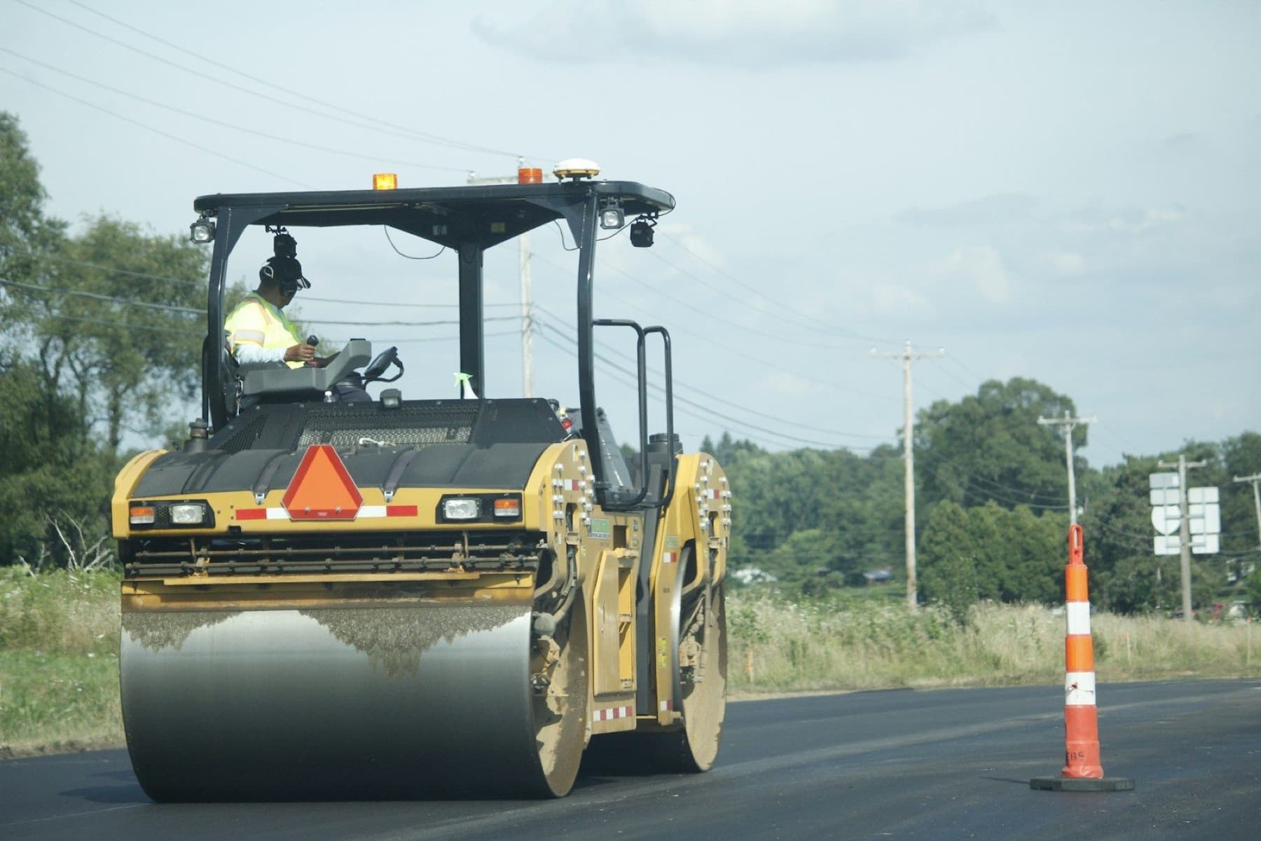 An operator in a safety vest drives a road roller over freshly laid asphalt beside a traffic cone