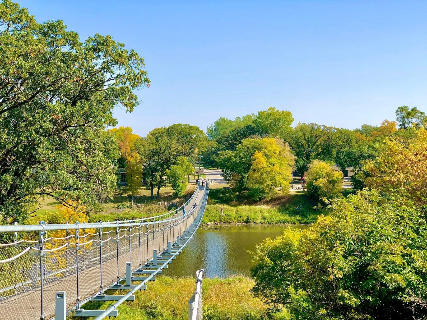 A pedestrian suspension bridge crosses a calm river between tree-lined park banks on a sunny day